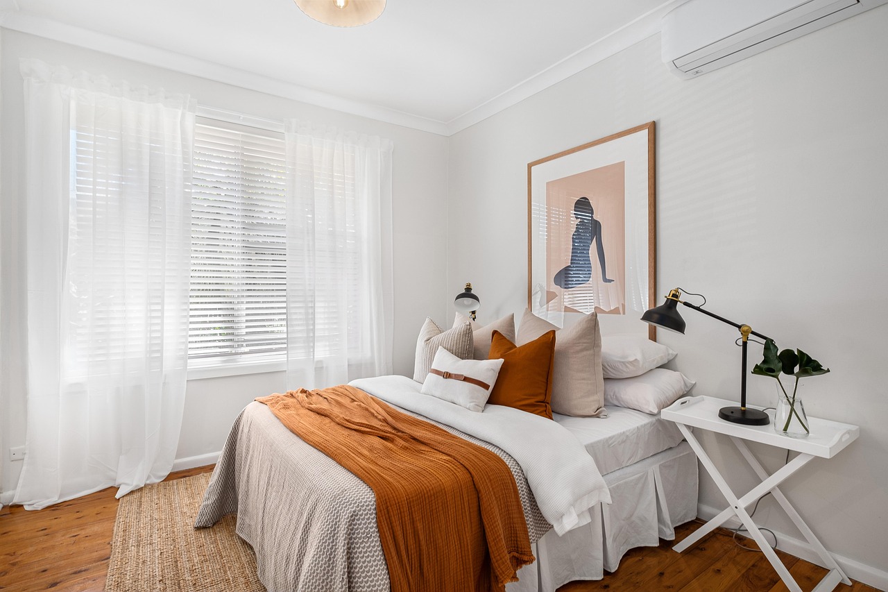 A cozy bedroom featuring a neatly made bed with decorative pillows, an orange throw blanket, and a minimalist bedside table with a lamp and a potted plant. Natural light streams through sheer curtains.